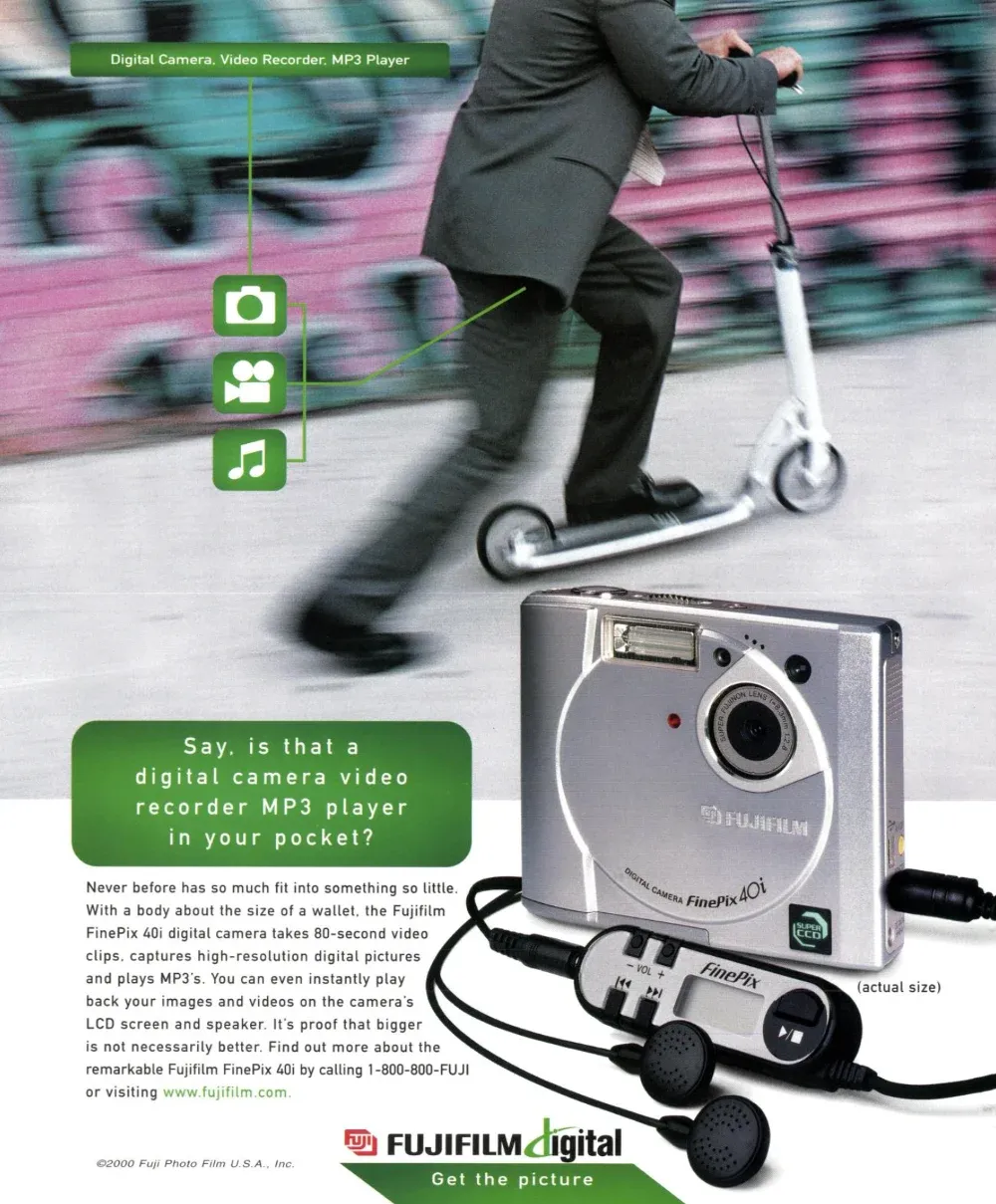 A suited man rides a kick scooter past a blurred wall while a Fujifilm FinePix 40i camera and its wired music remote fill the foreground.