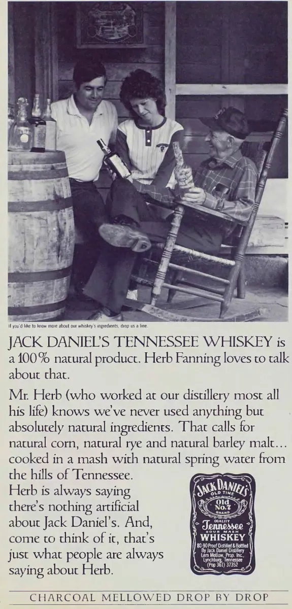 Herb Fanning sits in a wooden rocking chair holding an ear of corn while two visitors examine a whiskey bottle beside a barrel, above Jack Daniel's ingredients copy.