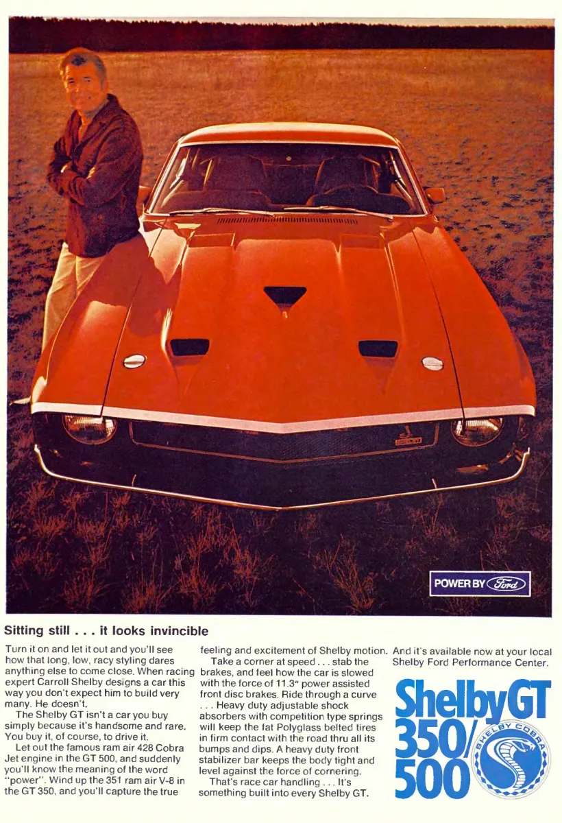 Carroll Shelby stands beside the front of an orange Shelby GT as the hood and grille fill most of the page.
