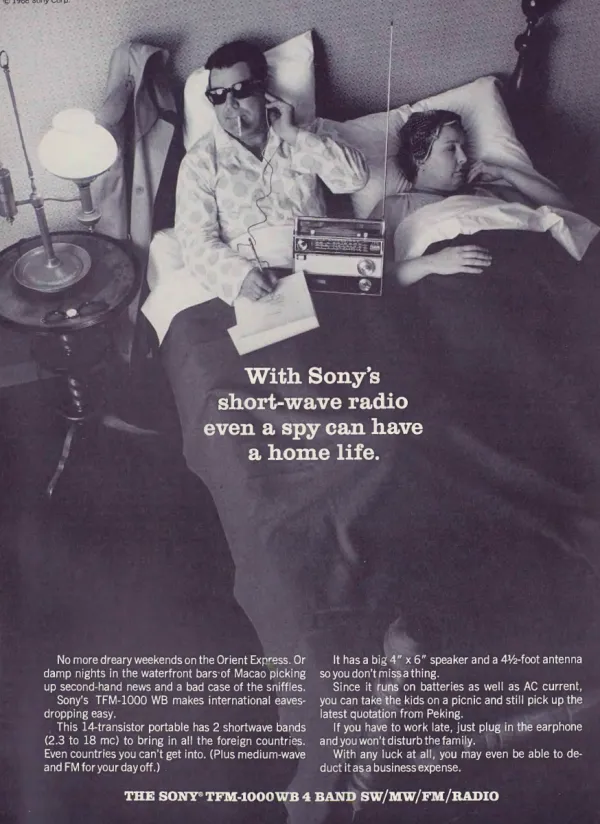 A man in sunglasses and pajamas listens to a Sony short-wave radio through an earphone and takes notes in bed beside his sleeping partner.