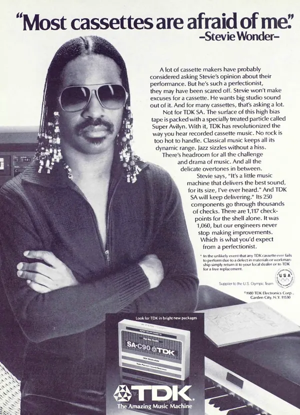 Stevie Wonder stands with folded arms beside a keyboard, beneath his cassette challenge, with a TDK SA-C90 package below.